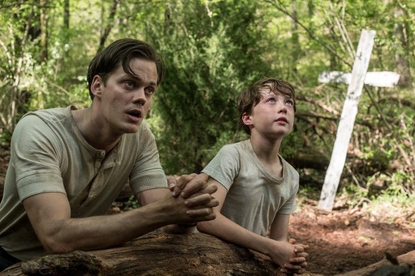 Bill Skarsgård dengan seorang aktor cilik memakai kaos lusuh sambil berdoa di sebuah kuburan dalam film The Devil All The Time